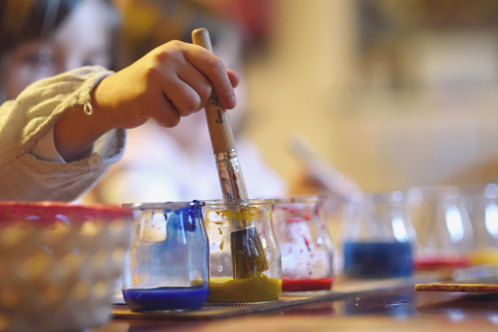 Criança segurando pincel com três vasos de tinta, representando atividades de artes manuais na pedagogia Waldorf.