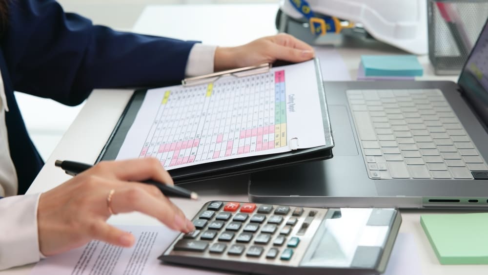 Controle de caixa escolar com planilha e calculadora ao lado de notebook durante conciliação bancária na secretaria.