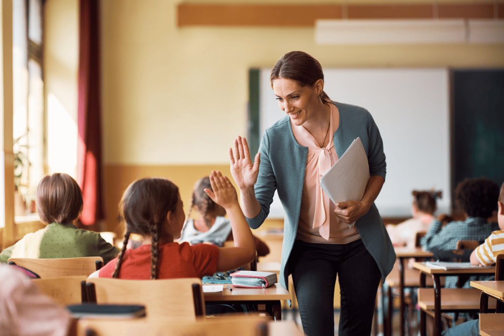 Professora dando um high five para aluna em sala de aula, destacando as regras na sala de aula de respeito e interação positiva.