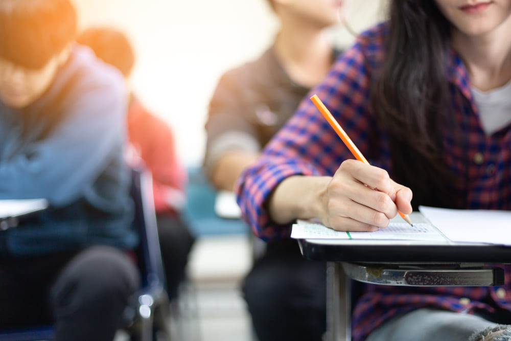 Estudante escrevendo em uma prova, com foco na mão segurando um lápis. A imagem remete à escolha de inglês ou espanhol no Enem, com foco na concentração durante a avaliação.