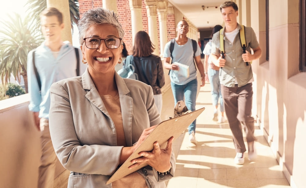 Diretora escolar sorrindo com prancheta, em um corredor escolar, no Dia do Diretor Escolar, com alunos ao fundo.
