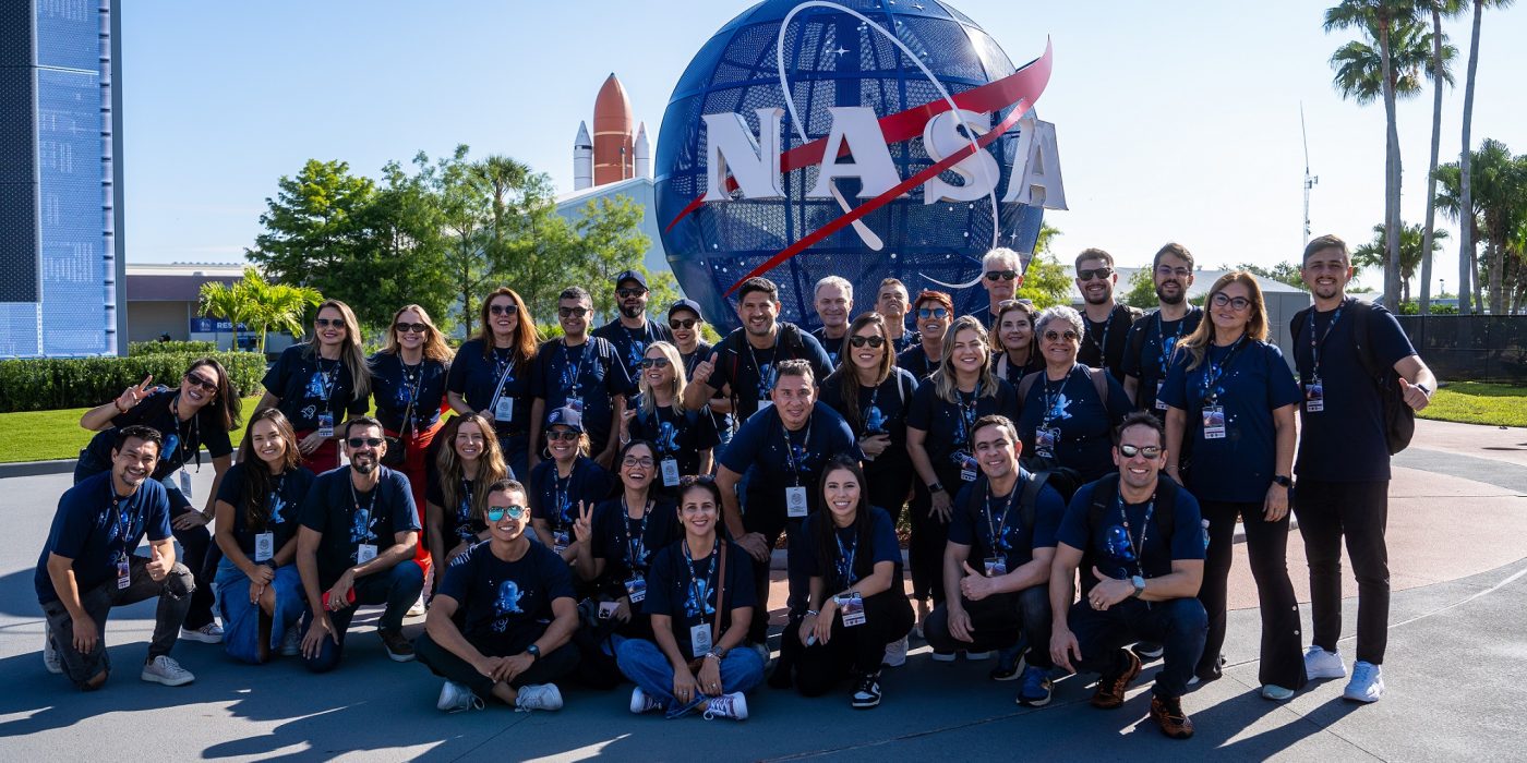 Estudantes e gestores escolares posam em grupo para foto em frente a logo da NASA no Kennedy Space Center