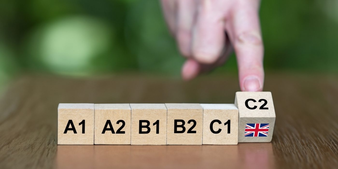 Blocos de madeira com os níveis de inglês A1, A2, B1, B2, C1 e C2, com uma mão apontando para o bloco C2.