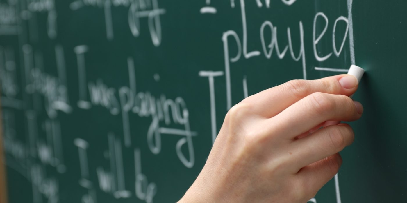 Mão escreve frases em inglês com giz branco em uma lousa verde durante aula de gramática em contexto bilíngue.