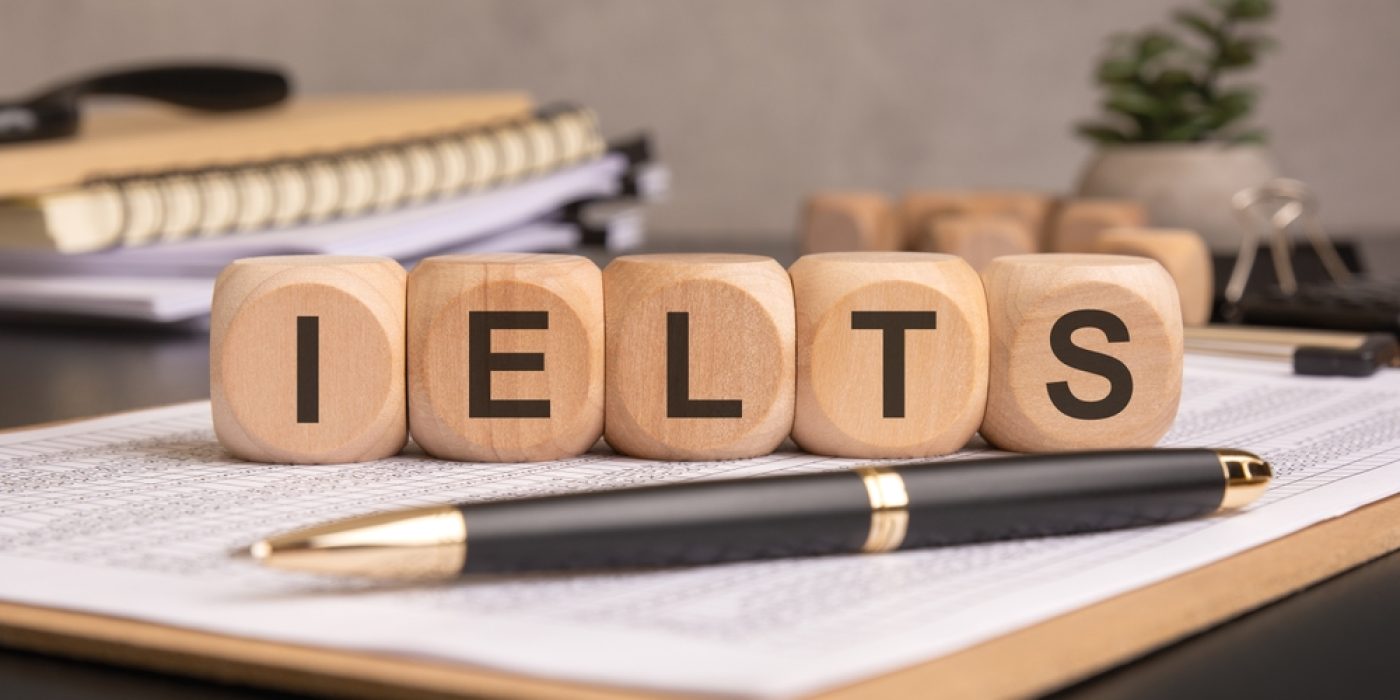 Blocos de madeira com as letras IELTS sobre uma mesa com documentos, caneta e caderno.