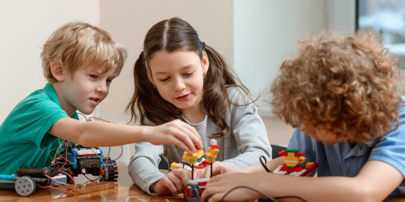 Dois meninos e uma menina, todos com cerca de 8 anos, brincam de montar um lego robotizado em cima de uma mesa de madeira.