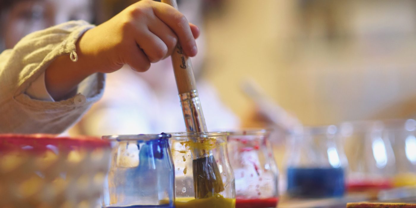 Criança segurando pincel com três vasos de tinta, representando atividades de artes manuais na pedagogia Waldorf.