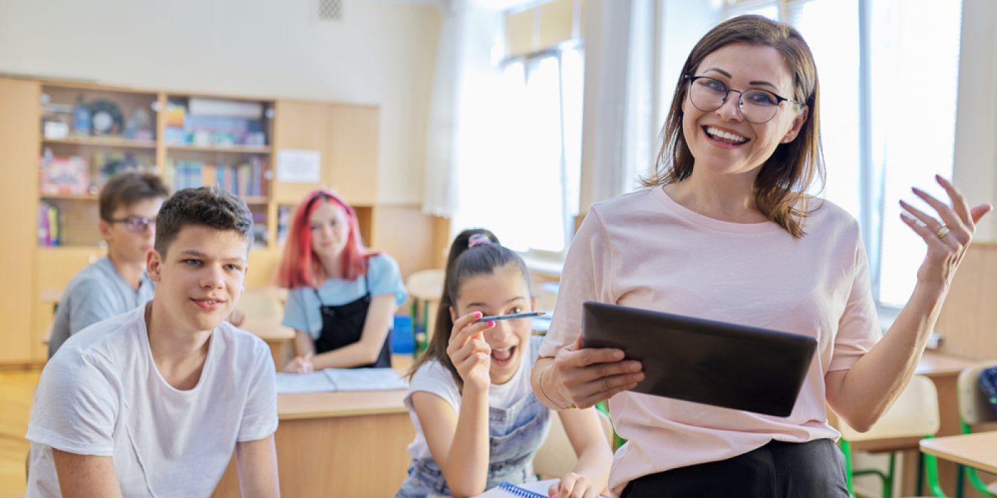 professora sorrindo com alunos adolescentes em sala de aula. Professora de pé e alunos sentados.