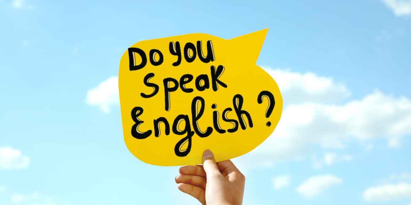Cartaz amarelo com a frase “Do you speak English?” sendo segurado por uma mão contra o céu azul.