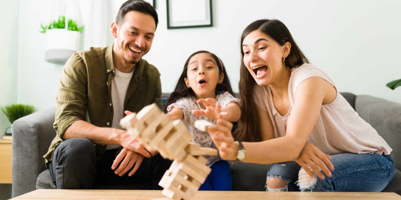 Pai, filha pequena e Mãe estão sentados no sofá e reagem com espanto e alegria ao ver que a torre de blocos de madeira que estão montando está começando a cair.