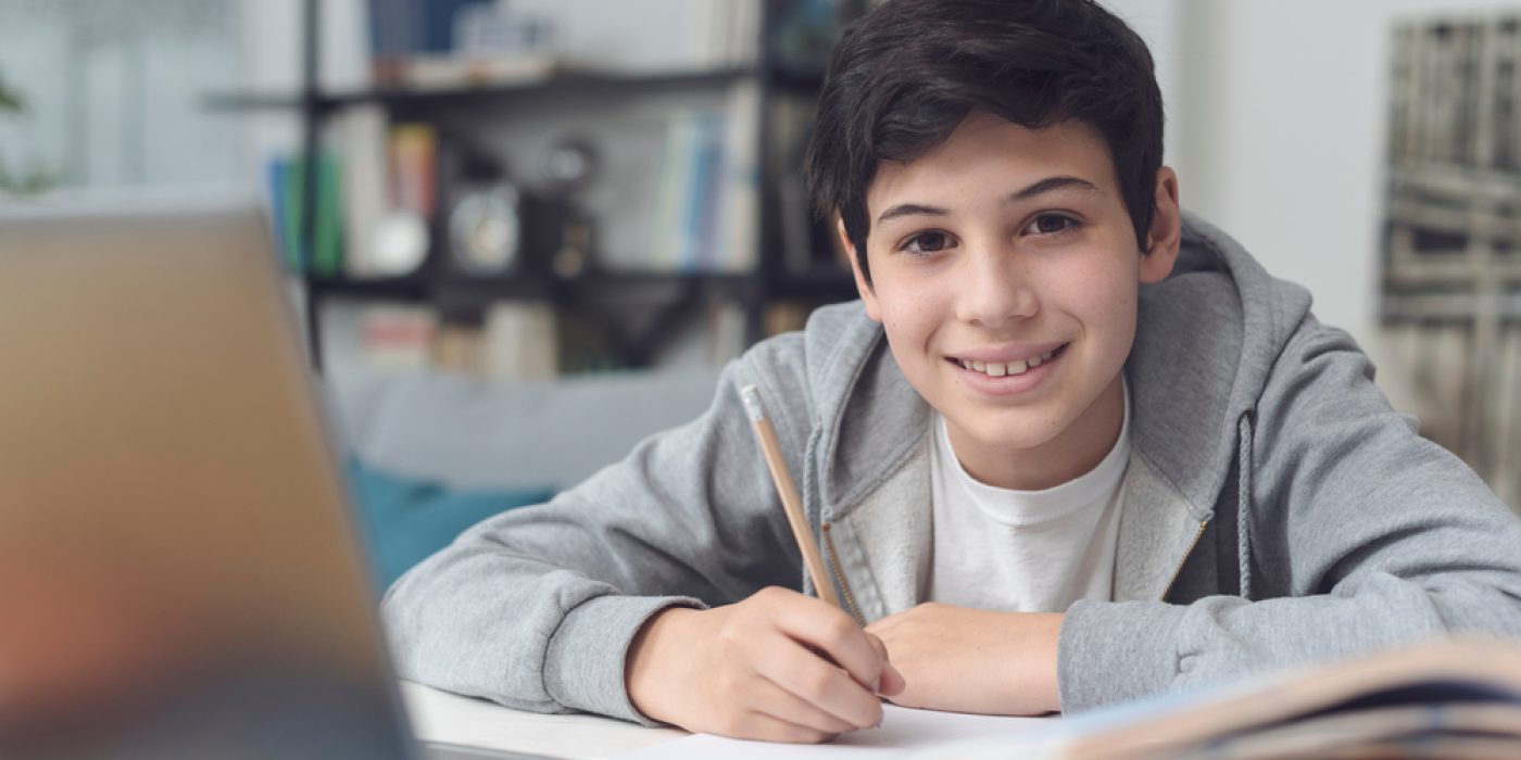 menino adolescente sentado, escrevendo em um caderno com um notebook à frente, sorrindo para a câmera