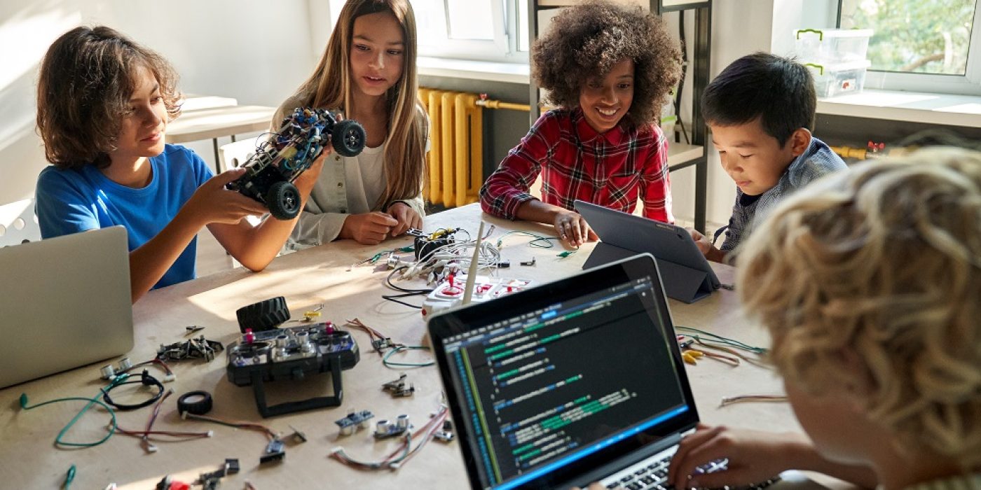 Crianças em atividade prática de programação e robótica, usando laptop, tablet e peças eletrônicas, representando o conceito de “pensamento computacional” no ensino fundamental.