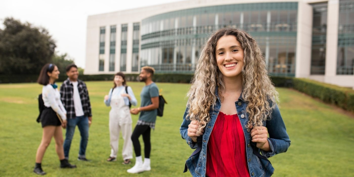 Jovem mulher segurando a alça da mochila e sorrindo para a câmera, com quatro estudantes conversando em grupo ao fundo, em um gramado de campus moderno.