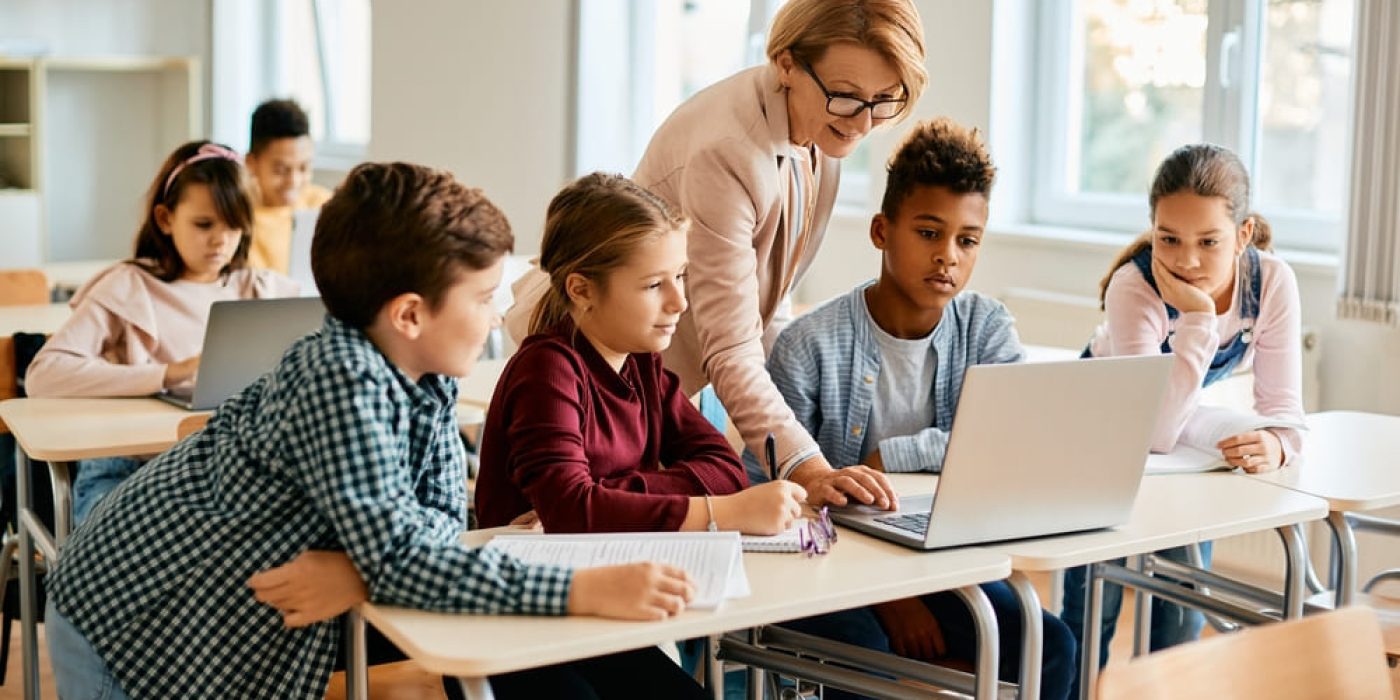 Professora orientando alunos no uso do computador em sala de aula, prática de letramento digital.
