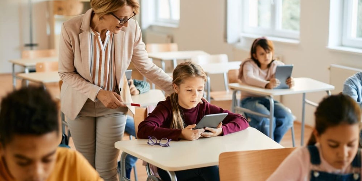 Professora ajudando aluna com tablet em sala de aula, exemplificando o uso da inteligência artificial na escola.