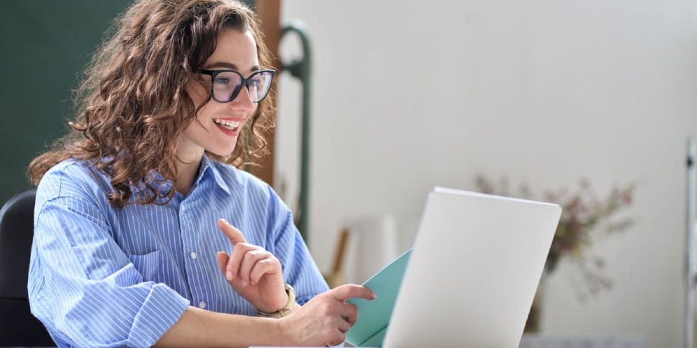 Mulher sorrindo em frente ao laptop, segurando um caderno e em ambiente tranquilo fazendo aulas de inglês para obter sua proficiência no idioma.