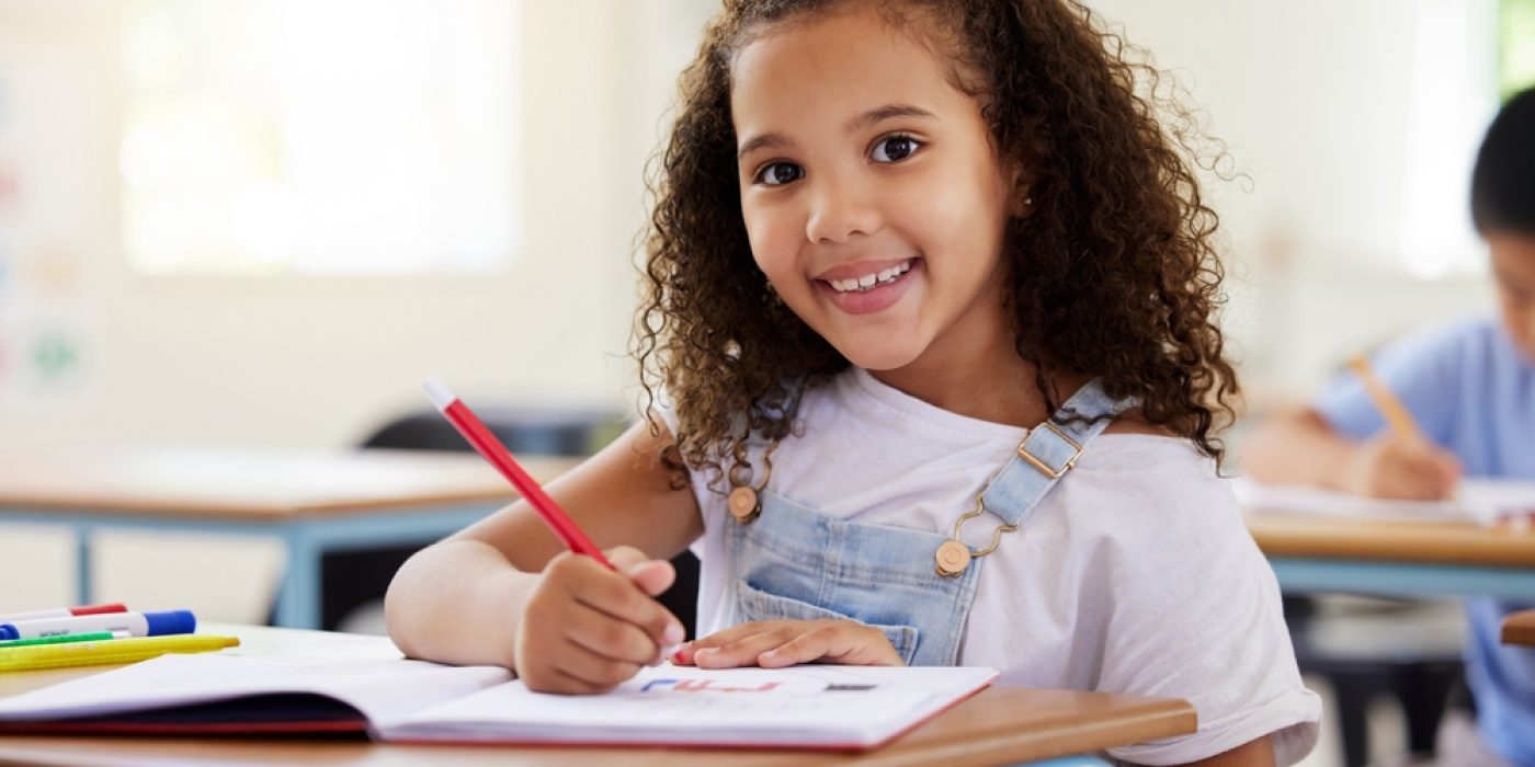 Menina de 6 anos escreve no caderno sentada na carteira da escola sorrindo para a câmera