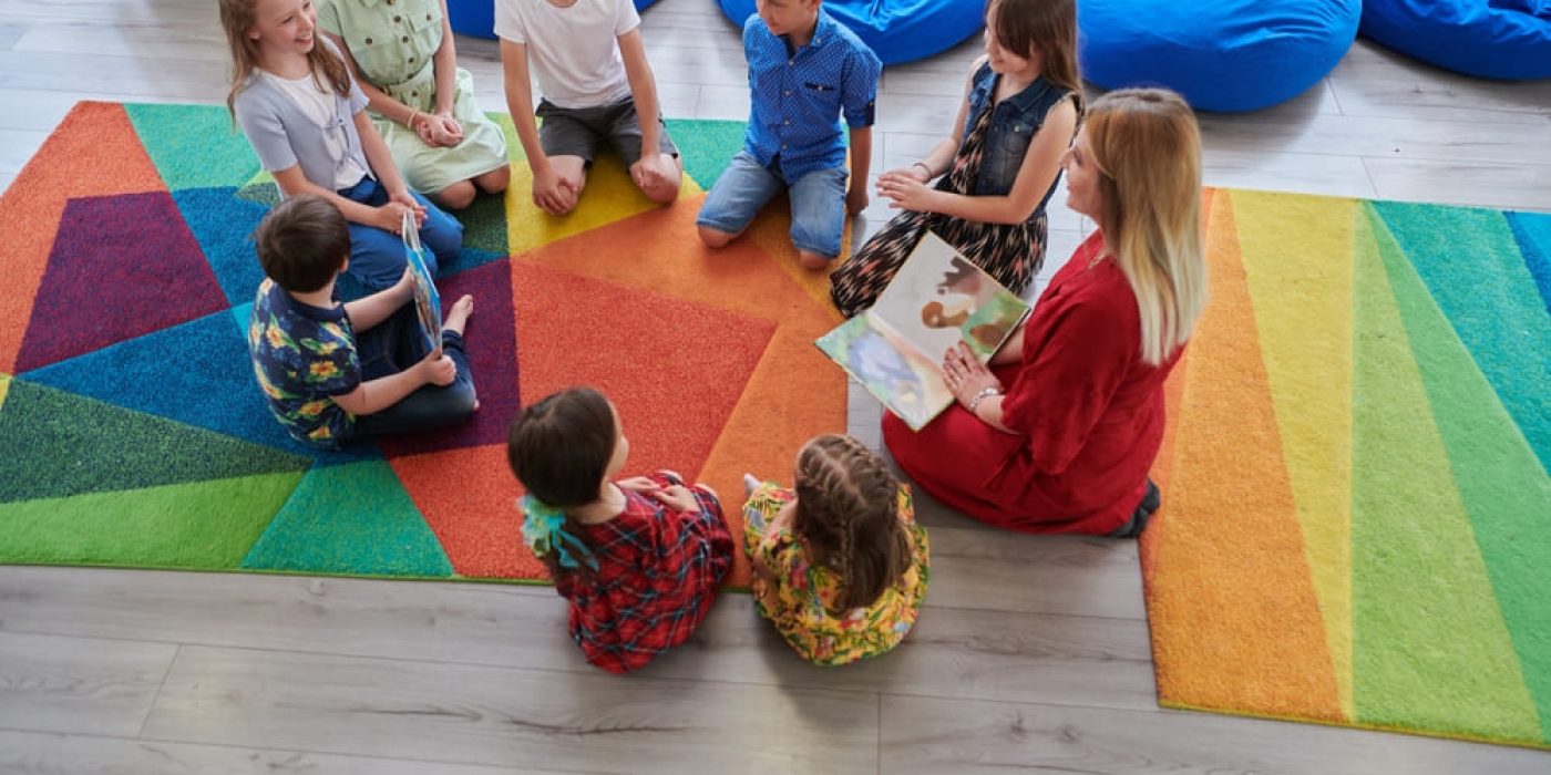 Professora lendo um livro para crianças em uma sala de aula infantil, com crianças sentadas ao redor em um tapete colorido.