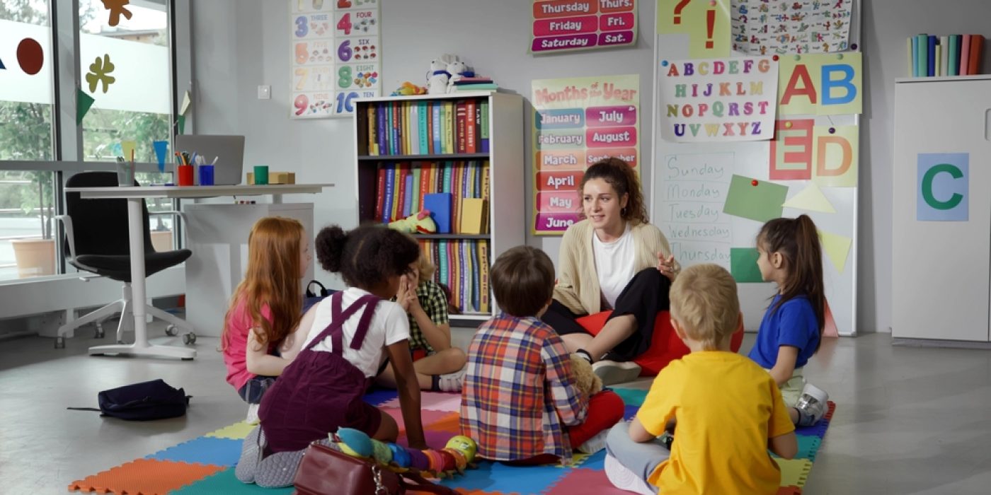 Crianças da Educação Infantil sentam em roda, no chão, junto com a professora.