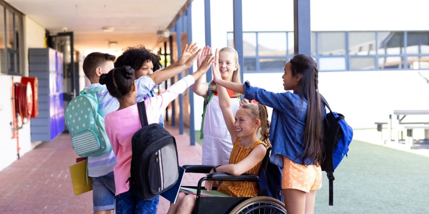 Crianças em corredor de escola celebrando juntas, incluindo uma aluna em cadeira de rodas, representando respeito às diferenças e inclusão escolar.