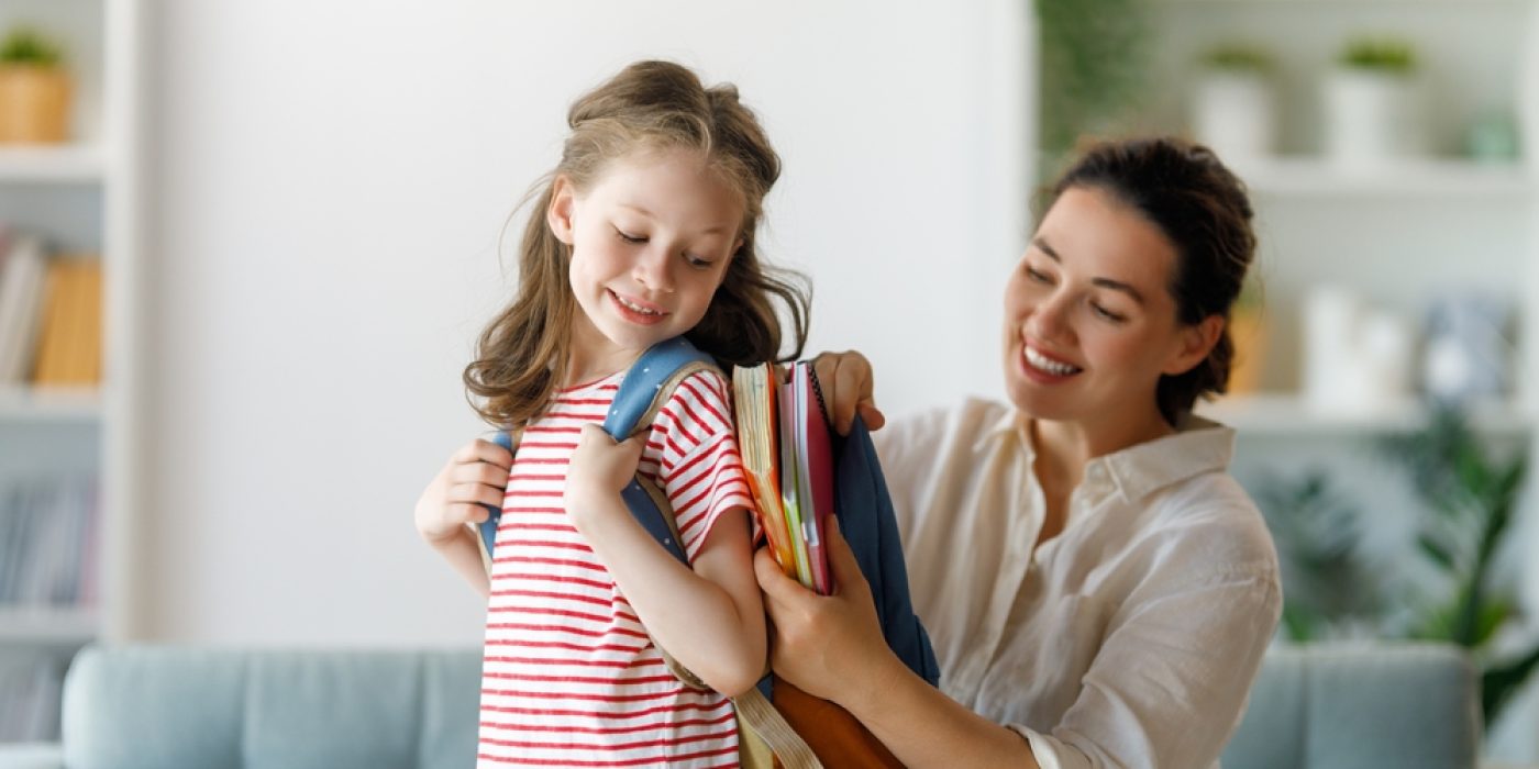 mãe ajuda filha pequena a colocar os materiais na mochila para ir à escola.