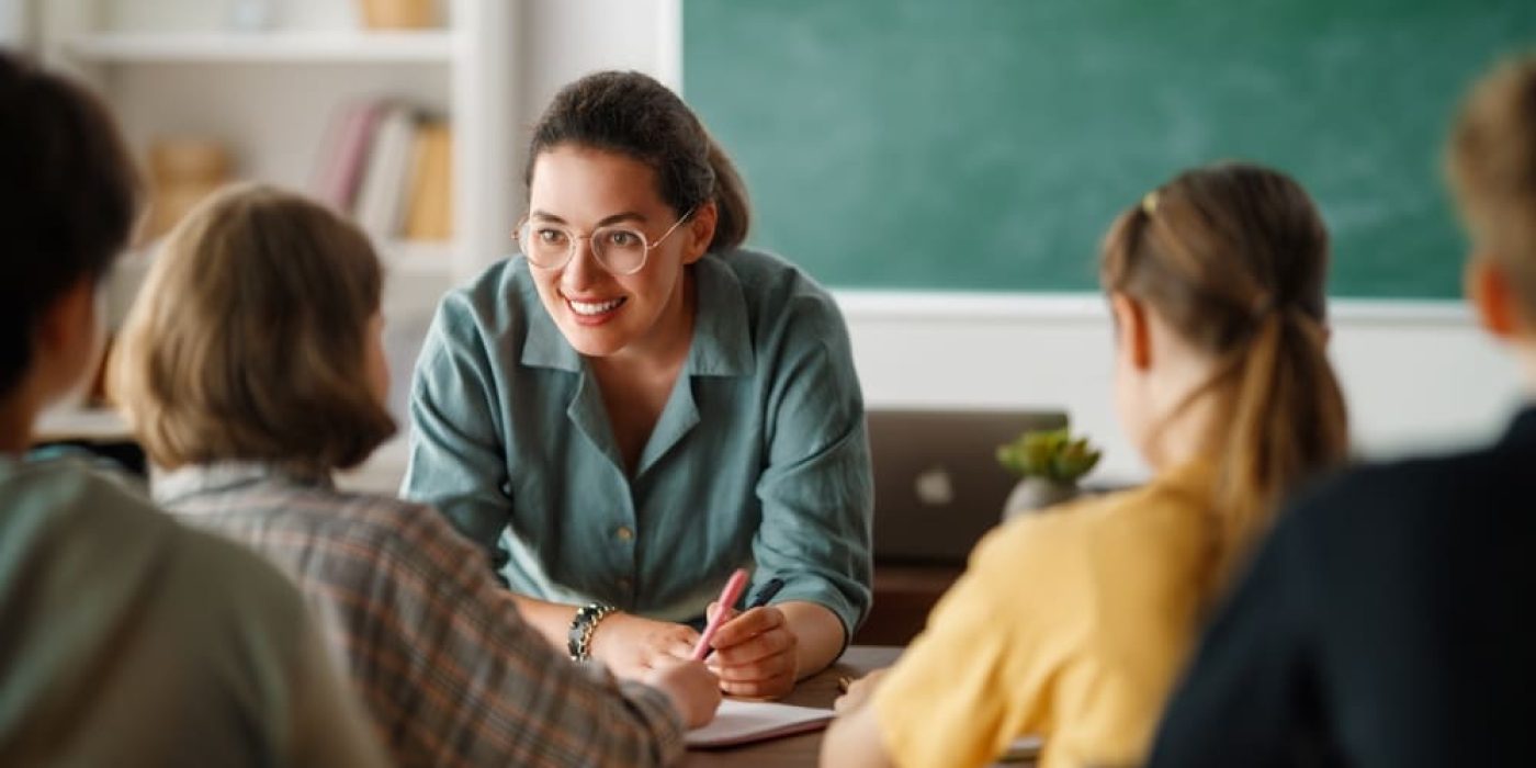 Professora na escola interagindo com seus alunos em sala de aula, com uma lousa verde ao fundo.