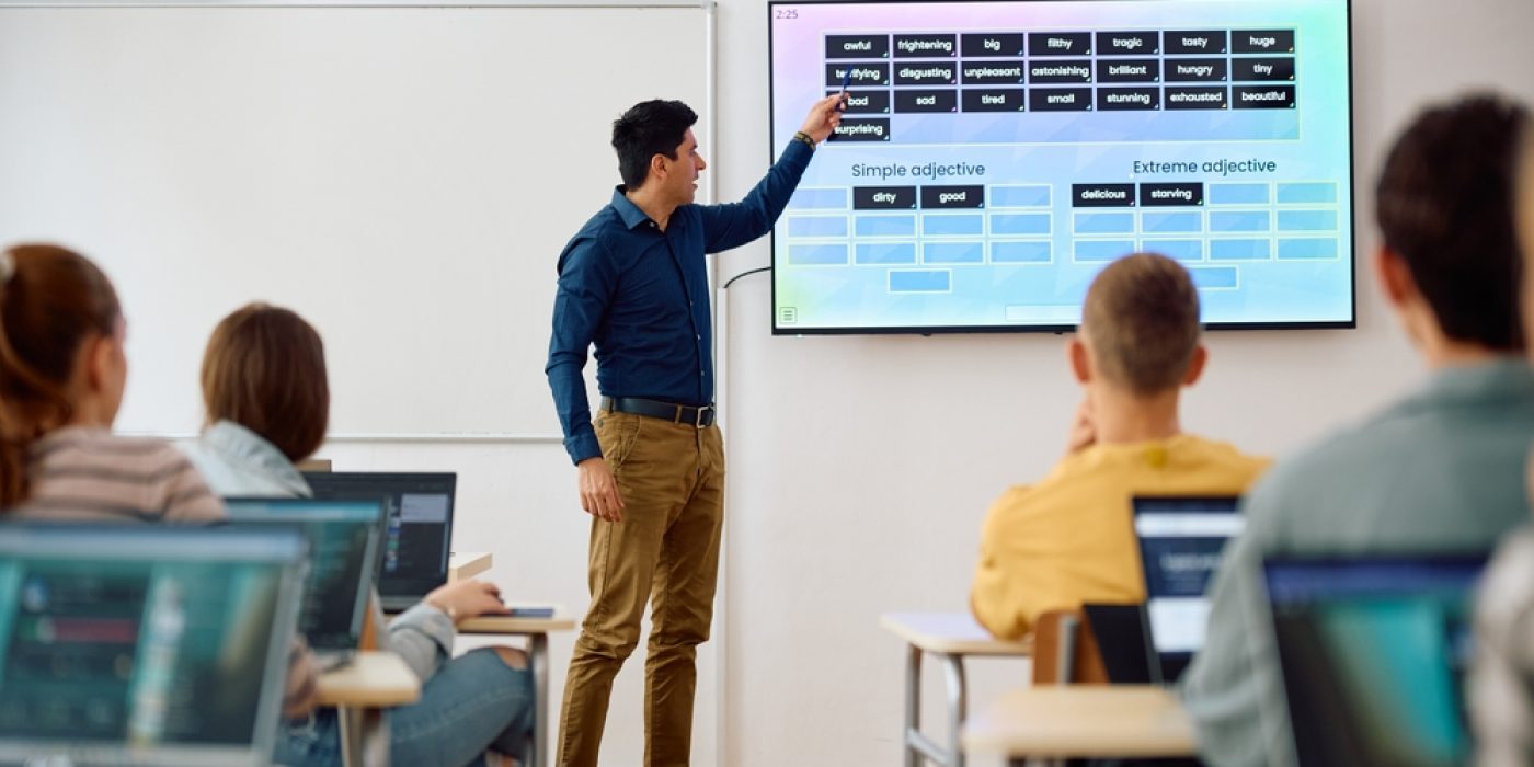 Professor apresenta conteúdo em sala de aula apontando para uma tela digital.