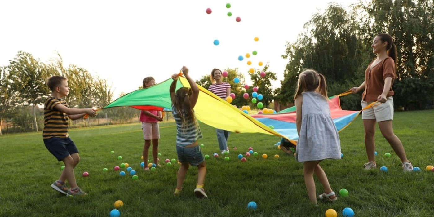 Crianças e professores brincando ao ar livre com uma lona colorida durante a Semana das Crianças, com bolinhas coloridas espalhadas pelo chão.