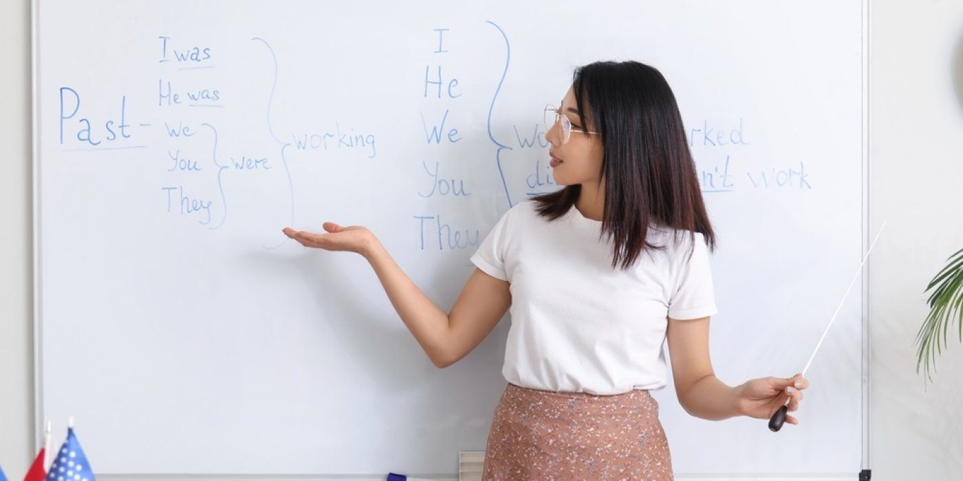 Professora ensina tempos verbais em inglês no quadro branco, ilustrando a importância da língua inglesa na comunicação global e na educação.