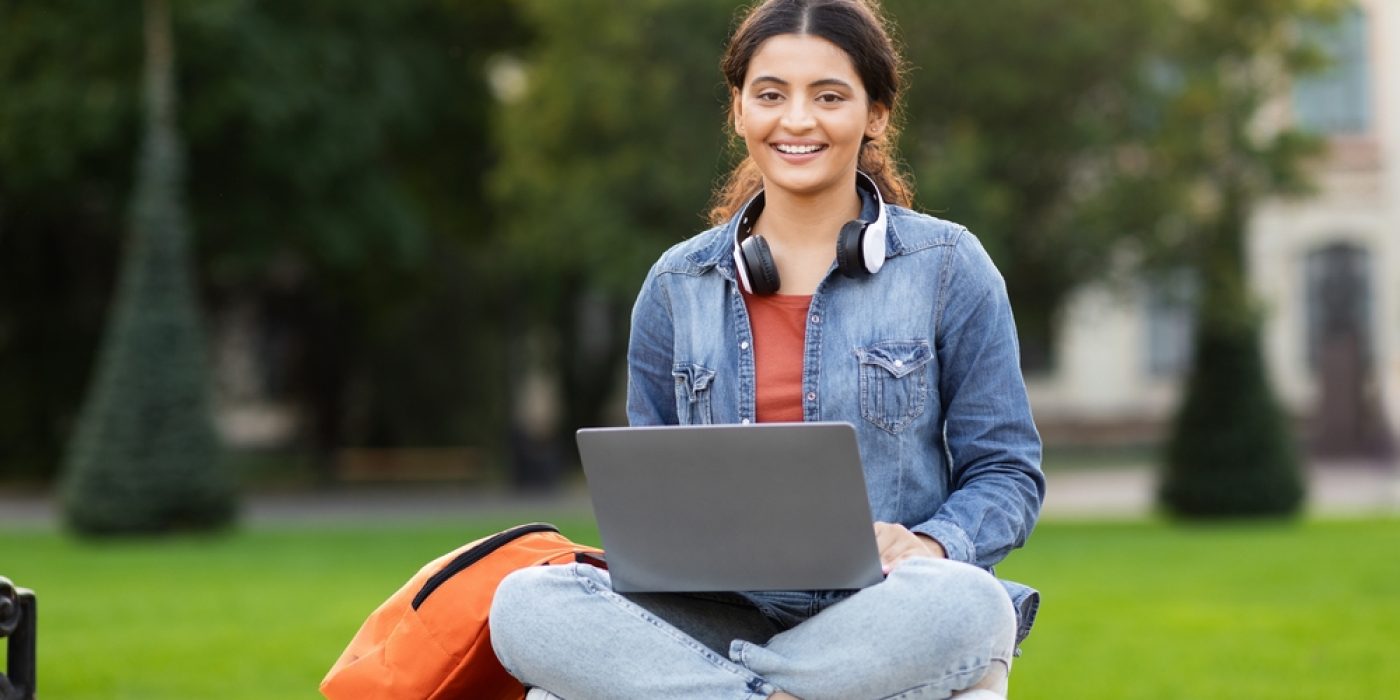 Estudante menina adolescente sentada ao ar livre sorri para a câmera com notebook no colo, headphones no pescoço e mochila ao lado.