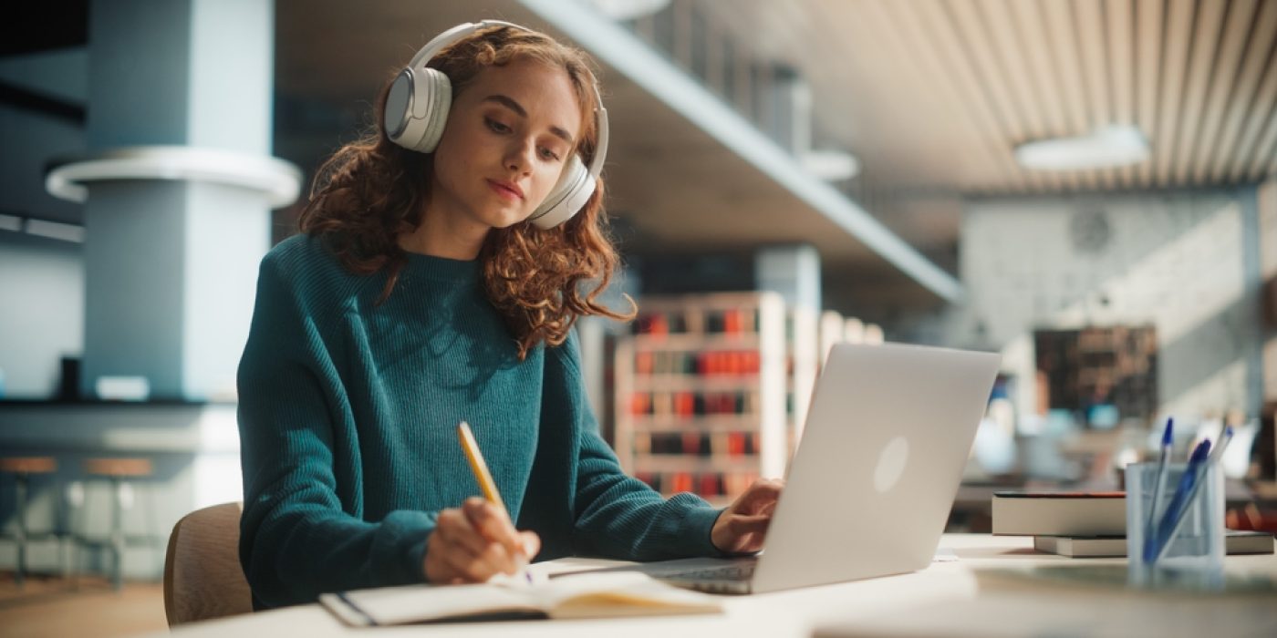 garota adolescente com cabelos ondulados e castanhos na altura dos ombros, veste moletom fechado verde escuro. Sentada na mesa de uma biblioteca, usa fones de ouvido brancos e escreve em um caderno enquanto usa o notebook.