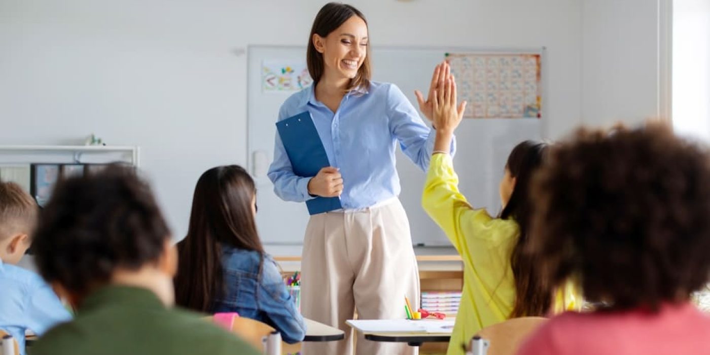 Professora sorridente cumprimenta aluno em sala de aula, representando o corpo docente escolar.