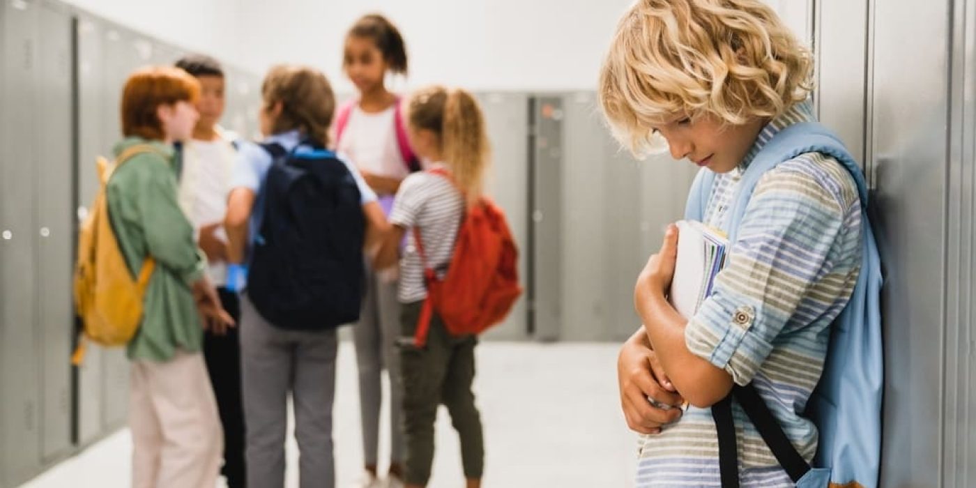 Menino isolado em corredor escolar enquanto colegas conversam em grupo, em cena que revela a importância de prevenir o bullying desde cedo na escola.