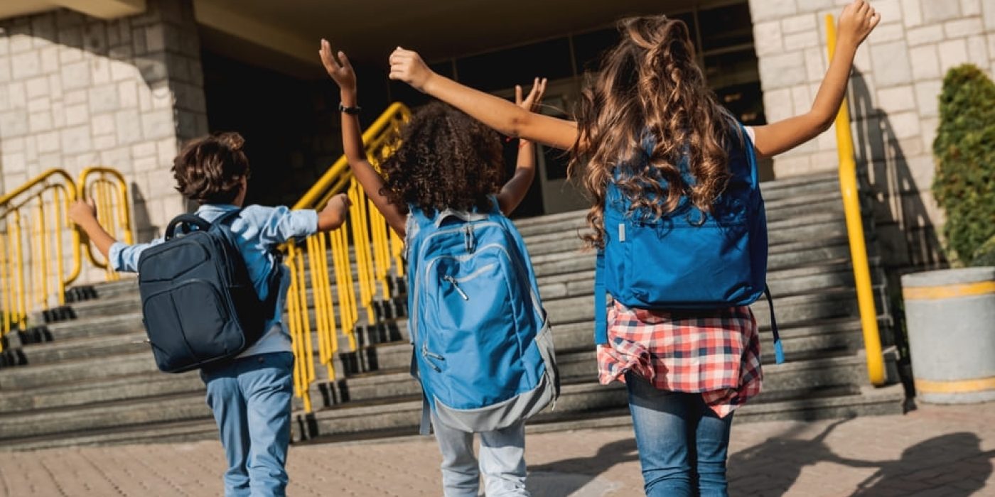 Crianças caminhando para a escola, com mochilas, comemorando o retorno às aulas.