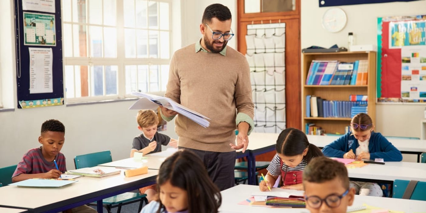 Professor acompanha alunos em atividades escritas em sala de aula, representando práticas relacionadas às tendências pedagógicas.