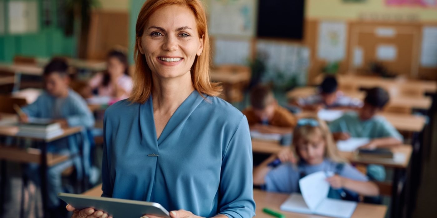 Professora segura um tablet em sala de aula, enquanto estudantes realizam atividades, representando a prática da avaliação formativa no acompanhamento da aprendizagem.