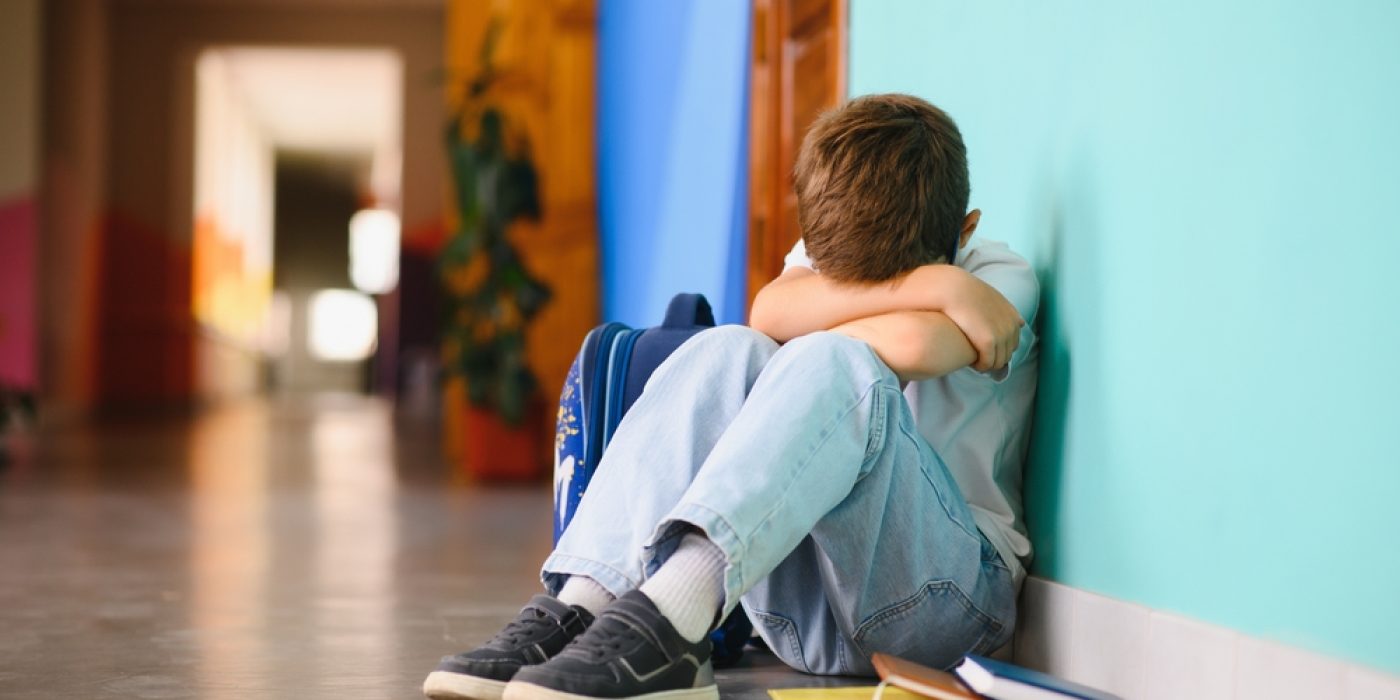 Menino de 7 anos sentado no corredor da escola com o rosto escondido nos braços.