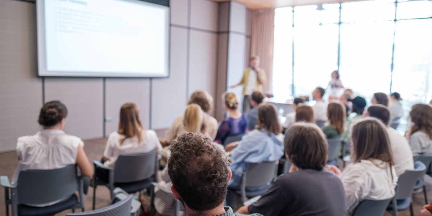 Pessoas assistindo a uma apresentação em sala de conferência, com foco em gestão pedagógica.