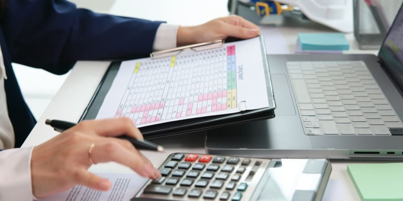 Controle de caixa escolar com planilha e calculadora ao lado de notebook durante conciliação bancária na secretaria.