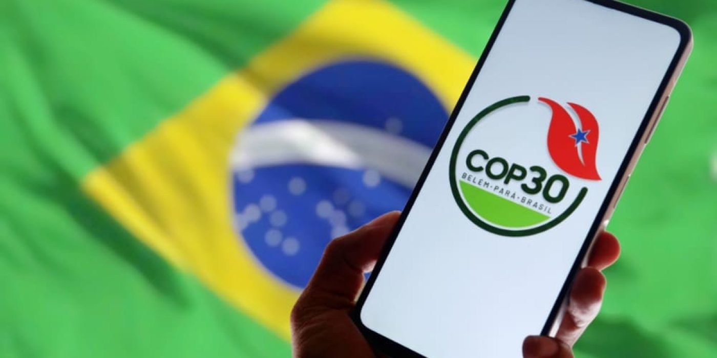 Logo da COP 30 no celular, com bandeira do Brasil ao fundo.