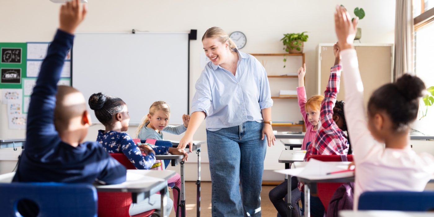Professora interagindo com crianças em sala de aula, enquanto vários alunos levantam a mão para participar.