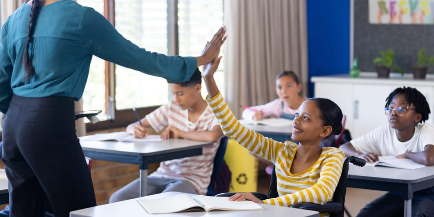 Criança em cadeira de rodas recebendo um high five da professora, exemplificando a inclusão através de tecnologia assistiva na educação.