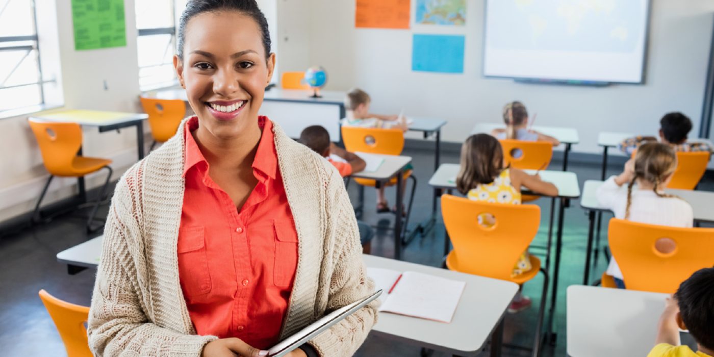 Professora negra de pele clara, cabelo preso, veste uma camisa salmão e cardigan bege claro. Ela sorri para a câmera, de pé, no meio da sala de aula, segurando um tablet nos braços. Ao fundo, crianças estão sentadas de costas nas carteiras.