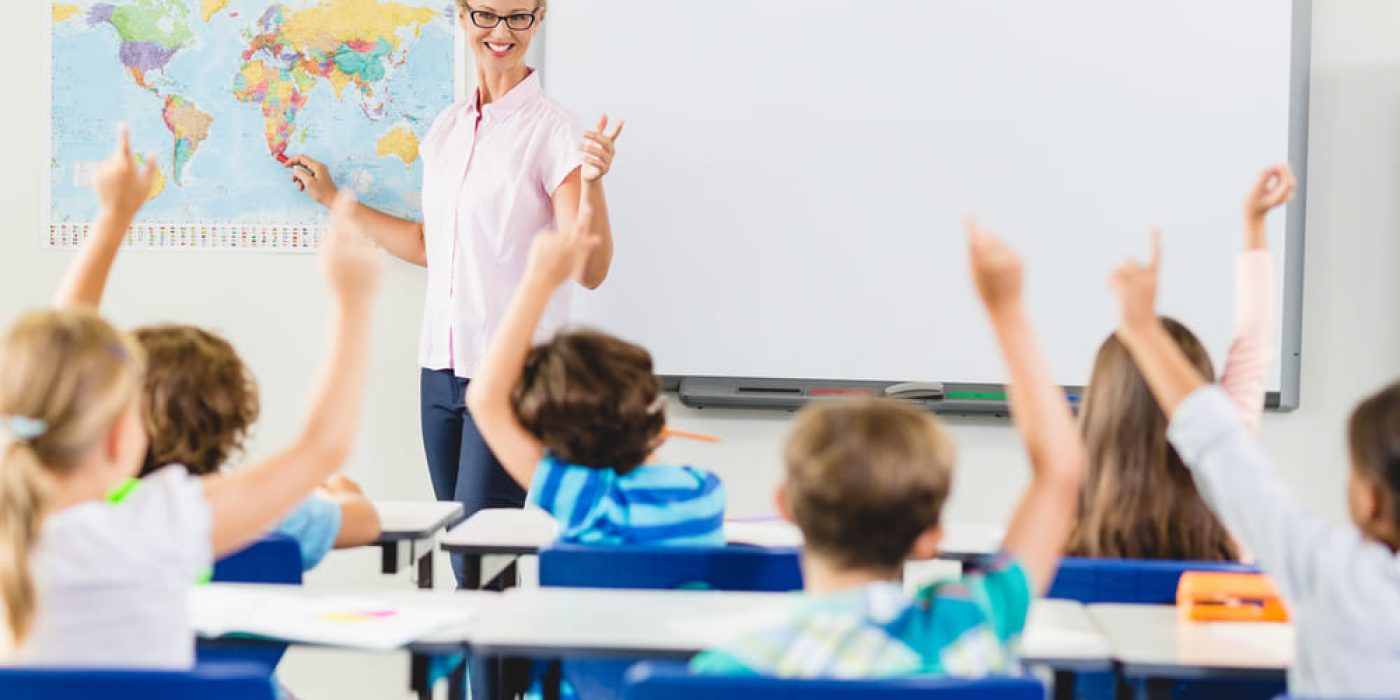 Professora aponta mapa; alunos levantam a mão em sala de aula, exemplificando uma abordagem pedagógica com participação.